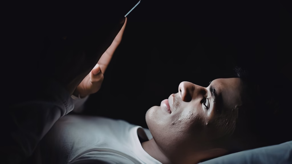 Man alone in bed at night, lit only by the glow of his phone screen.