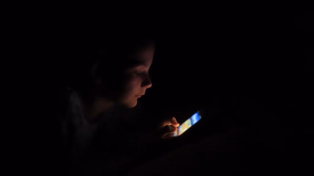 A child's face partially lit by the glow of a phone screen in a dark room